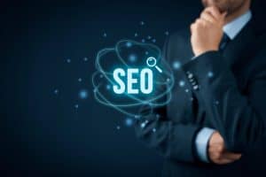 A person in a suit, thoughtfully holding their chin, stands next to the floating "SEO" text accompanied by a magnifying glass icon, symbolizing strategies on how to improve website SEO.